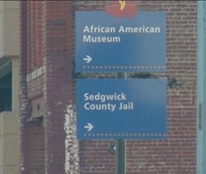 Két útmutató tábla egy oszlopban. Felső: 'African American Museum →' Alsó: 'Sedgwick County Jail →'